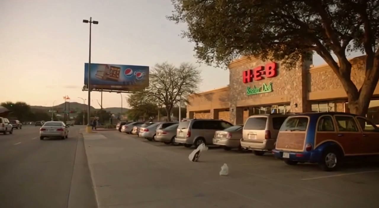 An H-E-B with a green Kosher Deli sign at dusk. A plastic bag blows across the lot past a wood-paneled PT Cruiser; a Pepsi billboard and the hills behind.